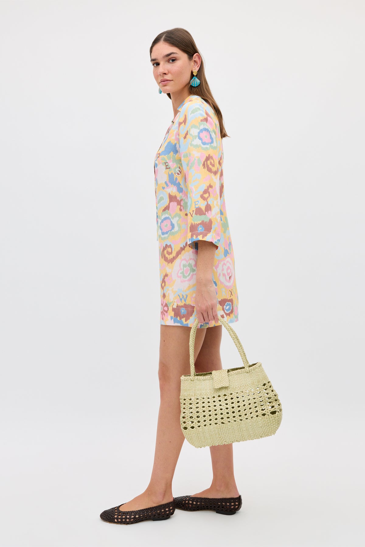 A woman in the Lucien - Ikat Bloom Yellow bohemian dress, black woven flats, blue earrings, and a light woven handbag stands against a plain white background, facing slightly to the side.