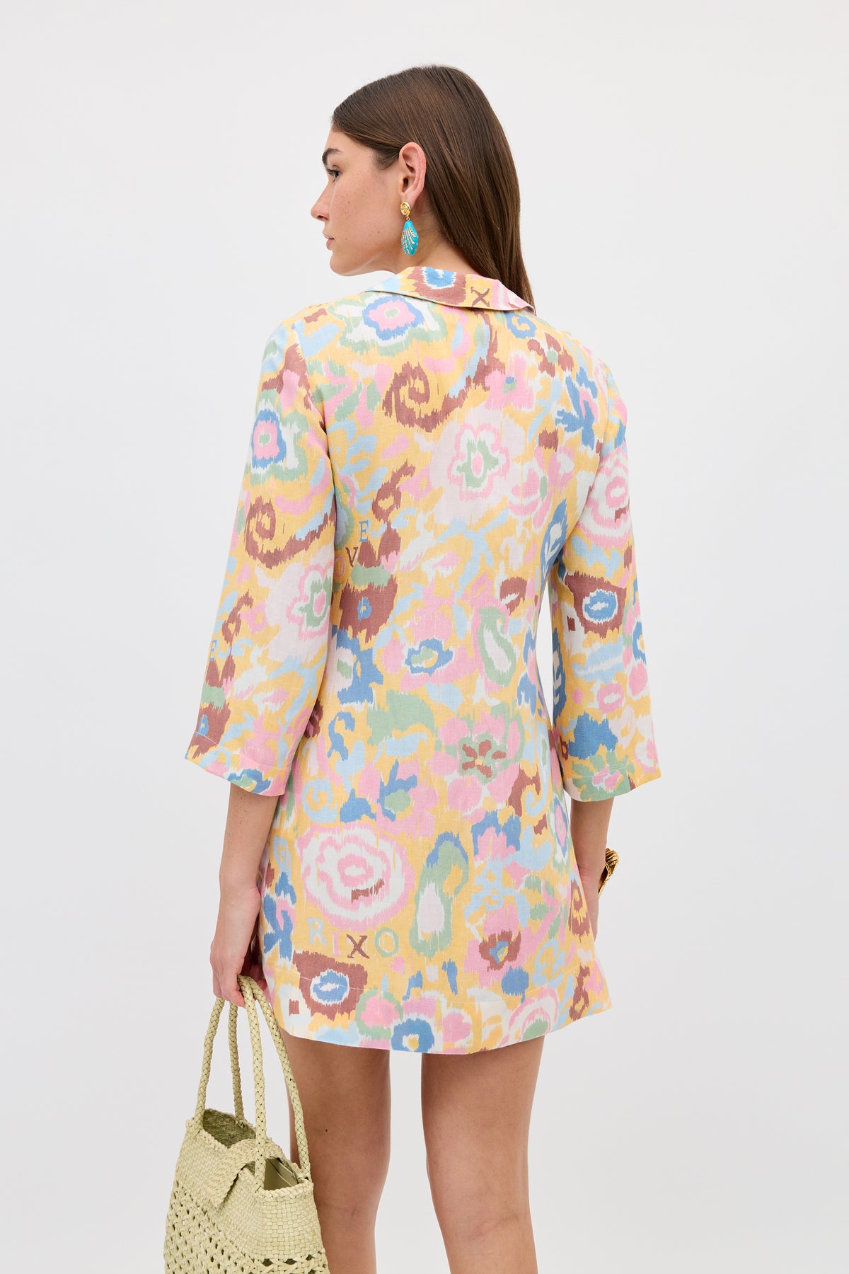 A woman with straight brown hair wears the Lucien - Ikat Bloom Yellow bohemian dress with three-quarter sleeves, carrying a woven straw bag and colorful earrings, facing away from the camera against a plain background.