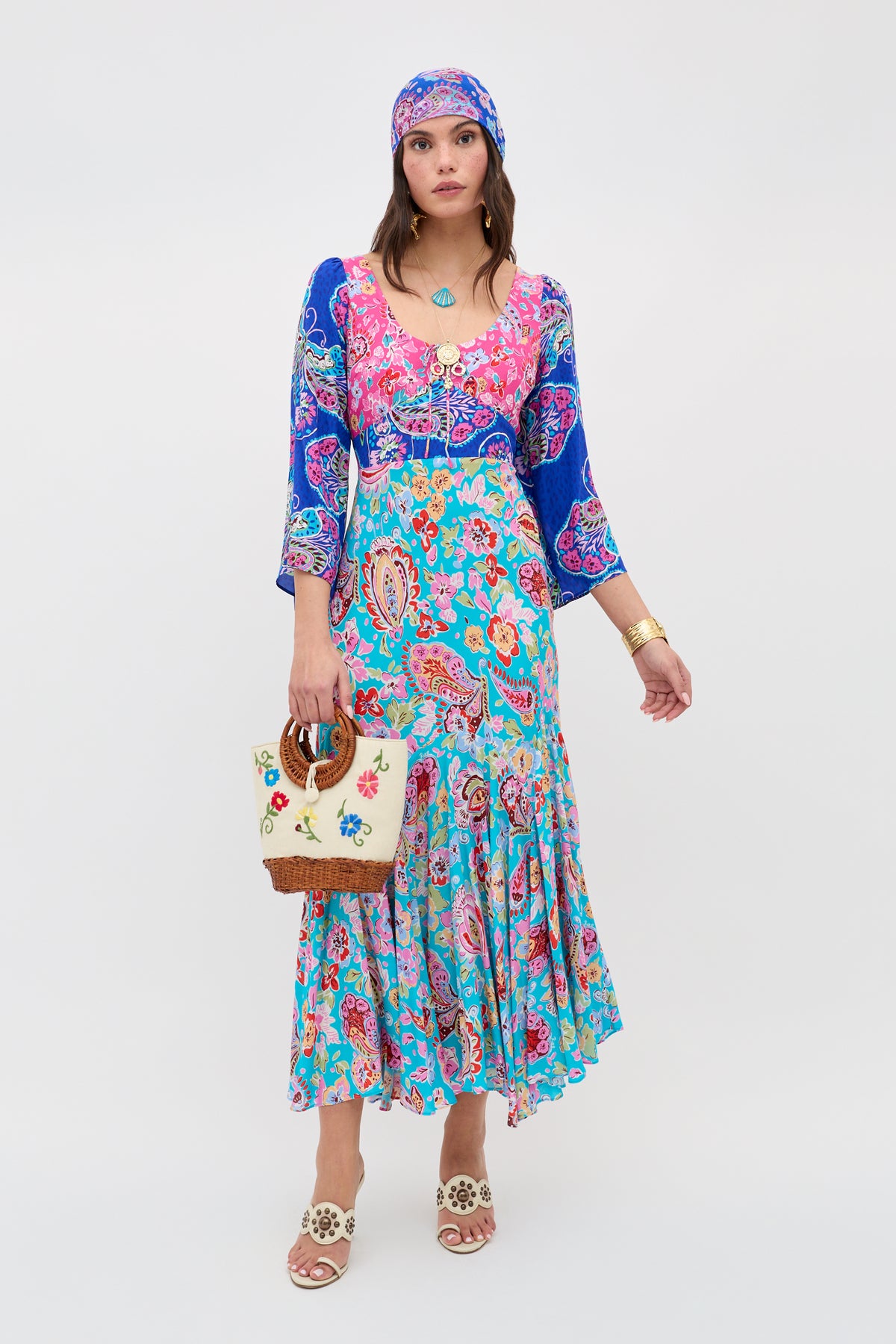 A woman wears the Karly - Cara Mix Blue dress, featuring colorful paisley patterns and three-quarter sleeves, paired with a matching headscarf, white sandals, necklace, gold bracelet, and a cream floral-detail handbag.