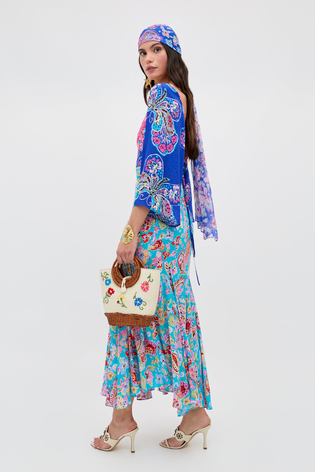 A woman in the Karly - Cara Mix Blue bohemian outfit with paisley and floral patterns, a matching headscarf, a cream embroidered handbag with wooden handles, and beige heeled sandals stands against a white background.