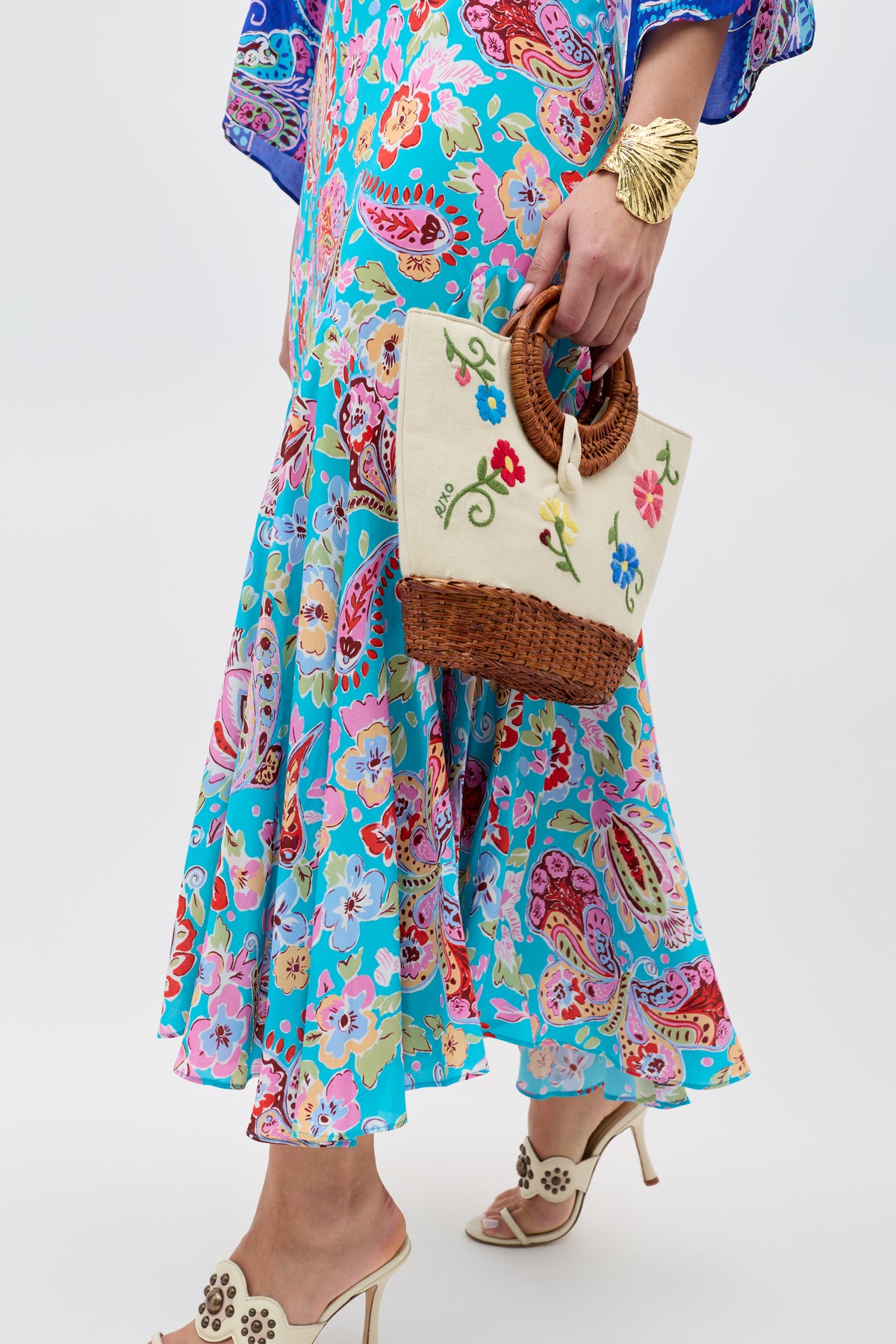A person in the Karly - Cara Mix Blue dress, white high-heeled sandals, and a gold bracelet holds a small woven handbag with floral embroidery and round handles. The background is plain light gray.