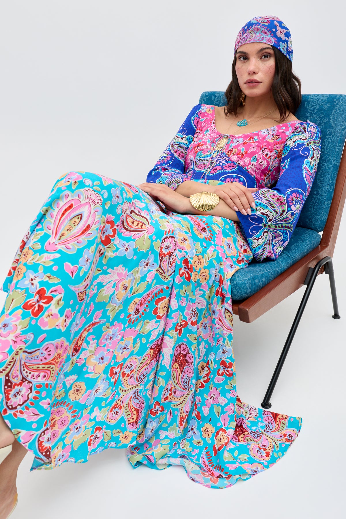 A woman wearing the Karly - Cara Mix Blue dress and matching headscarf sits on a blue upholstered chair, gazing confidently at the camera with a relaxed expression.