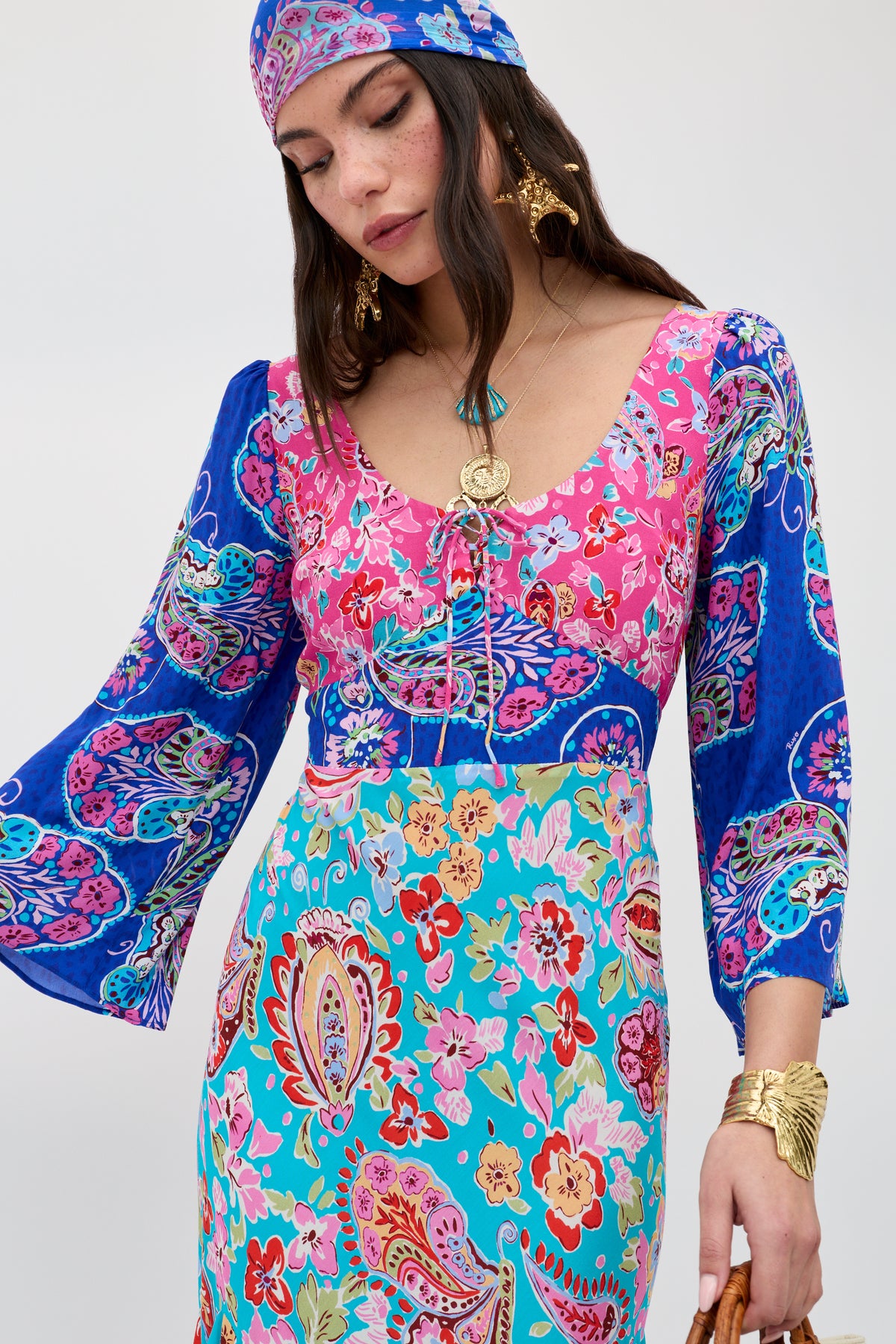 A woman wears the Karly - Cara Mix Blue dress, featuring colorful floral and paisley patterns with flared sleeves, gold jewelry, and a matching headscarf, gazing down serenely against a plain background.