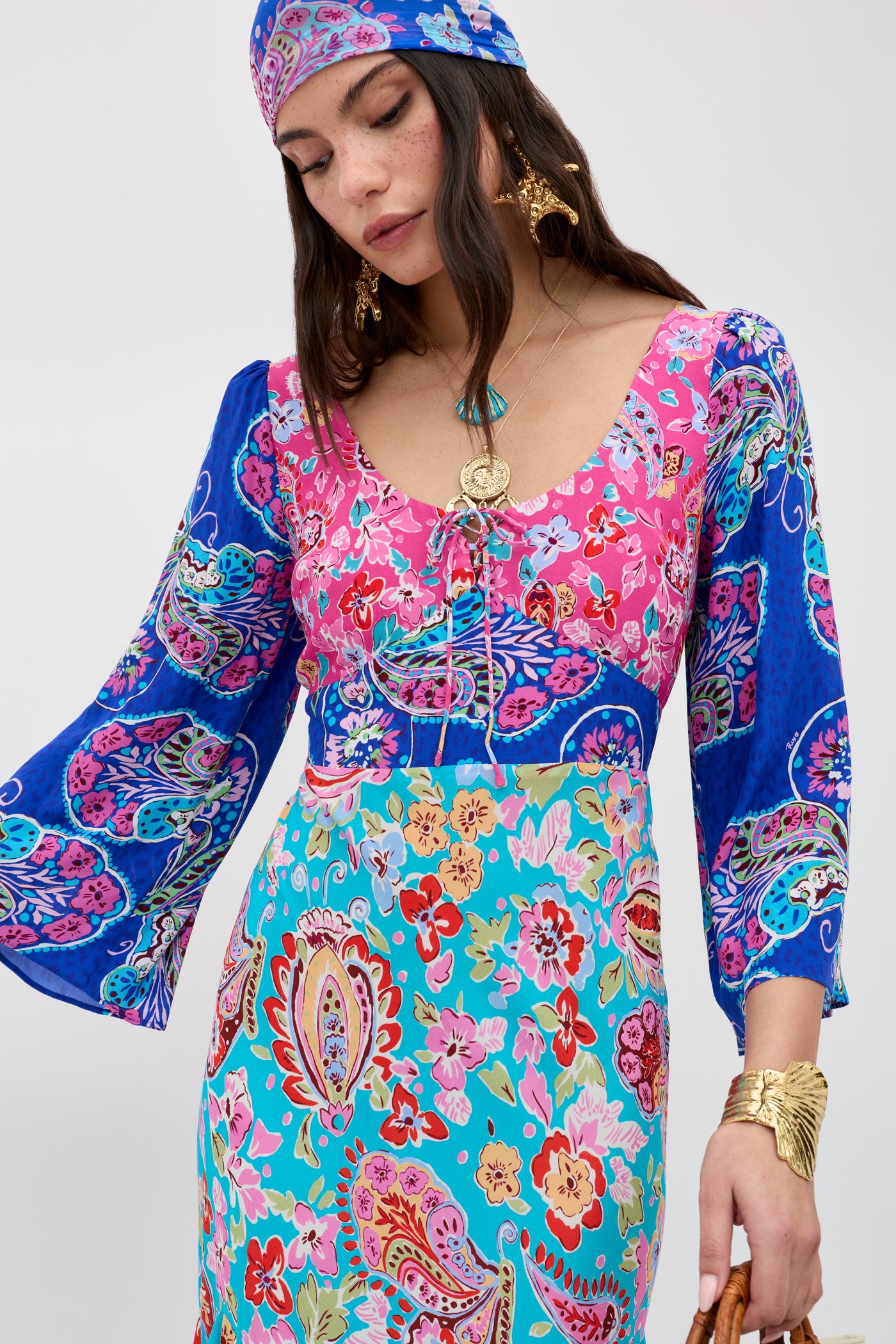 A woman wears the Karly - Cara Mix Blue dress, featuring colorful floral and paisley patterns with flared sleeves, gold jewelry, and a matching headscarf, gazing down serenely against a plain background.