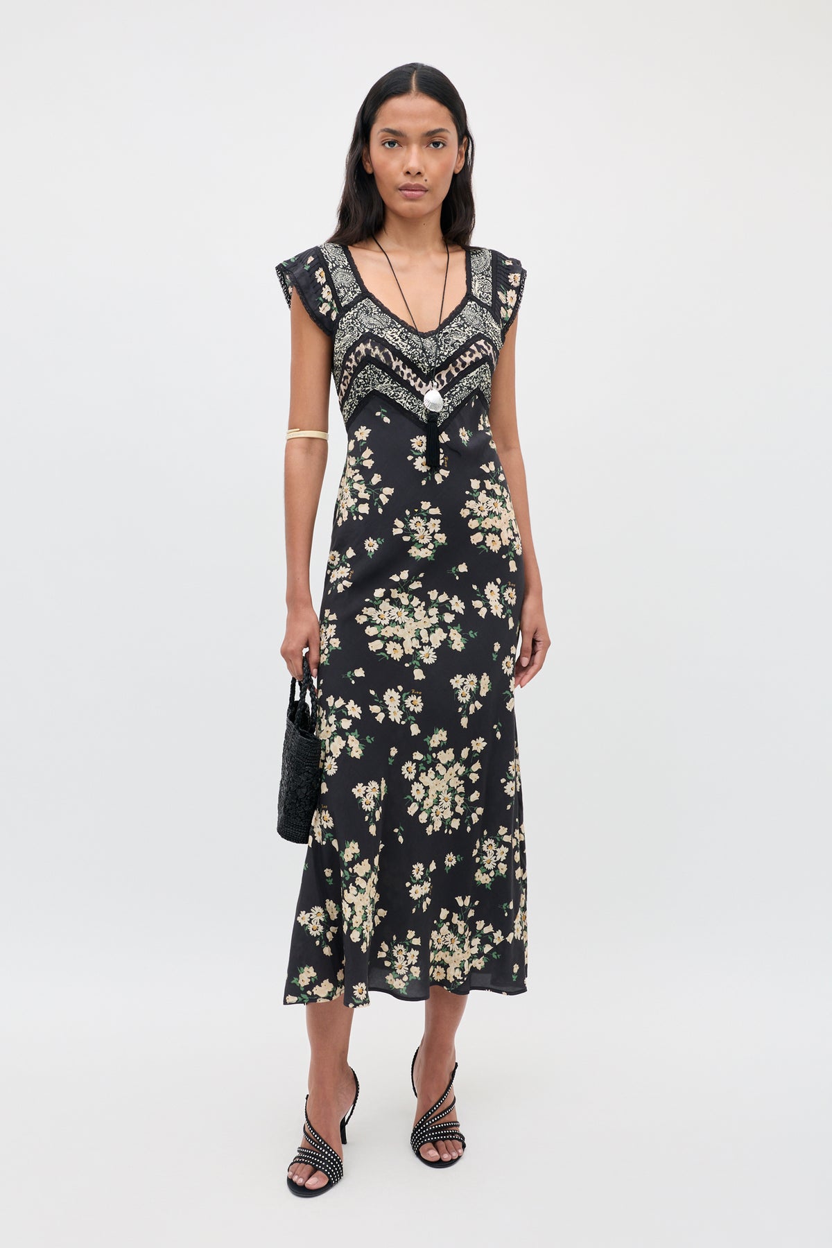 A woman stands against a plain background wearing the Bailee - Amelia Gremlin Mix Black dress, which features a black base with a floral pattern and decorative trim. She pairs it with black heeled sandals, a black handbag, jewelry, and a gold arm cuff. Her long hair is styled down and she has a neutral expression.