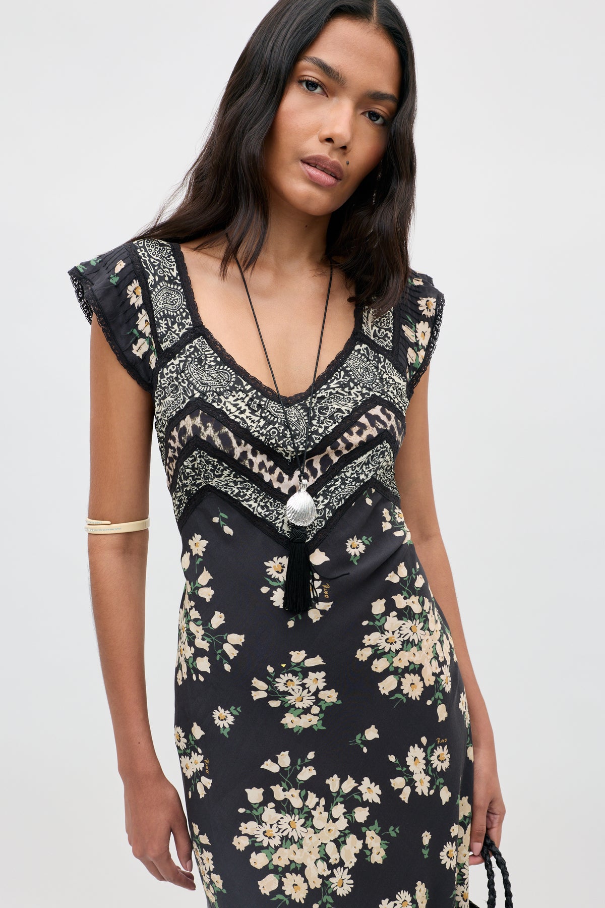 Wearing the Bailee - Amelia Gremlin Mix Black dress, a woman with long dark hair stands against a light background, accessorized with a pendant necklace and an arm cuff. The dress features striking floral and geometric patterns in black.