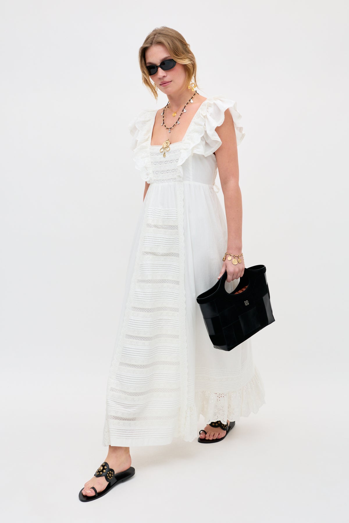 A woman wearing the Delaney - White long dress with ruffled sleeves, accessorized with sunglasses, black sandals, gold jewelry, and a black handbag, stands against a plain white background.