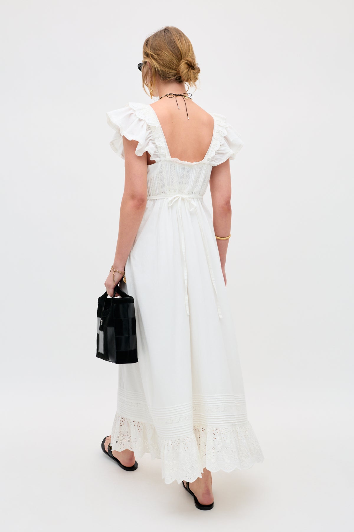 A woman with light hair in a loose bun wears the Delaney - White dress, featuring ruffled sleeves and a lace-trimmed hem. She holds a black handbag and stands facing away on a plain light background, paired with sandals and sunglasses.