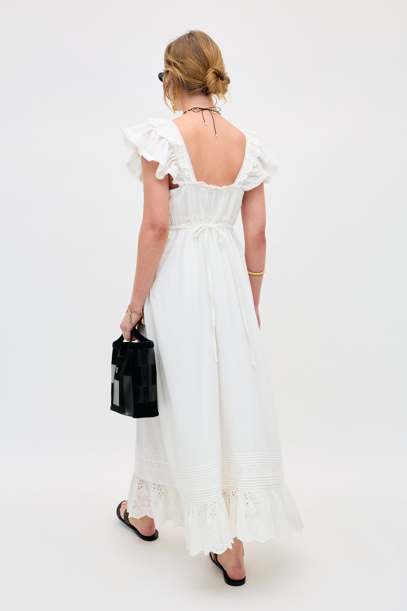 A woman with light hair in a loose bun wears the Delaney - White dress, featuring ruffled sleeves and a lace-trimmed hem. She holds a black handbag and stands facing away on a plain light background, paired with sandals and sunglasses.