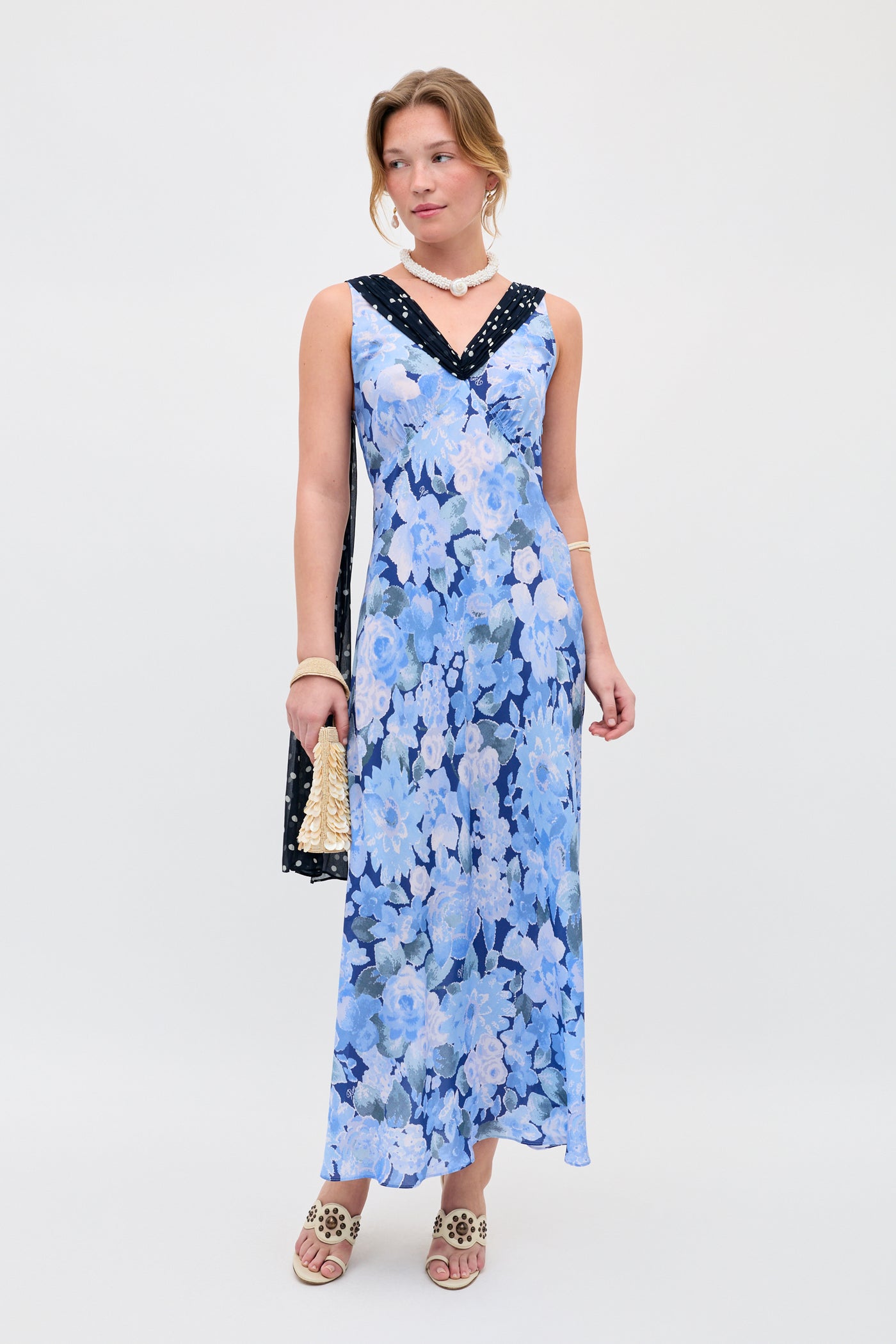 A woman stands against a plain background wearing the Roux - Chelsea Dot Mix Blue sleeveless, ankle-length dress with black neckline detail. She accessorizes with a beige purse, sandals, a bracelet, and a necklace.