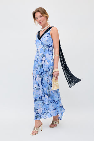 A woman stands against a plain light background wearing the Roux - Chelsea Dot Mix Blue sleeveless floral dress, paired with a black polka-dot scarf, beige floral-detail heels, gold bracelet, chunky necklace, and holding a small gold purse.
