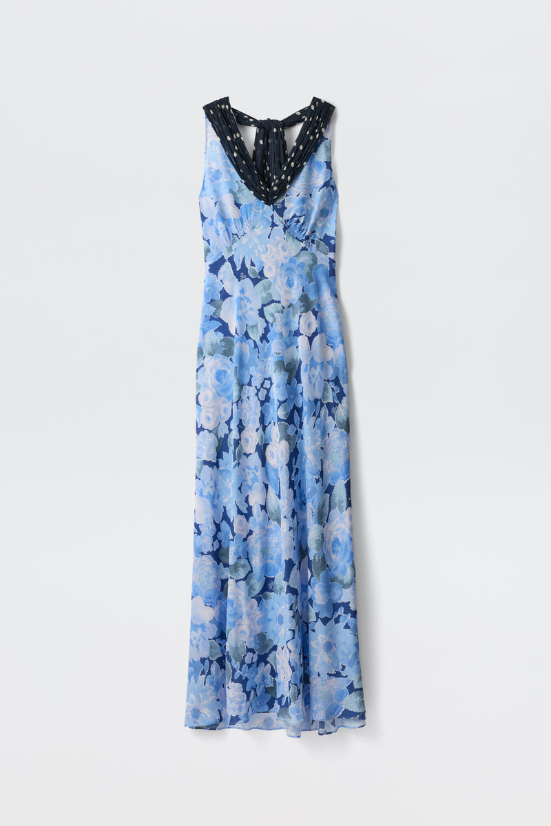 The Roux - Chelsea Dot Mix Blue is a sleeveless, floor-length dress featuring a blue floral pattern, black V-neckline with white dots, bow tie at the back, and flowing lightweight fabric.