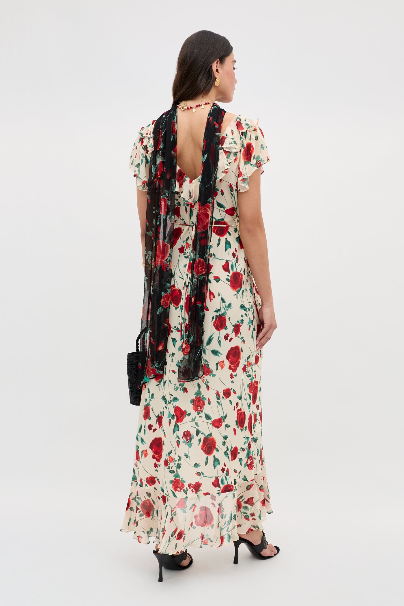 A woman wears the Meadow - Claudia Rose Cream dress, featuring a long cream silhouette with a red floral pattern. She pairs it with black heels, a lace scarf, and a black handbag against a plain white background.