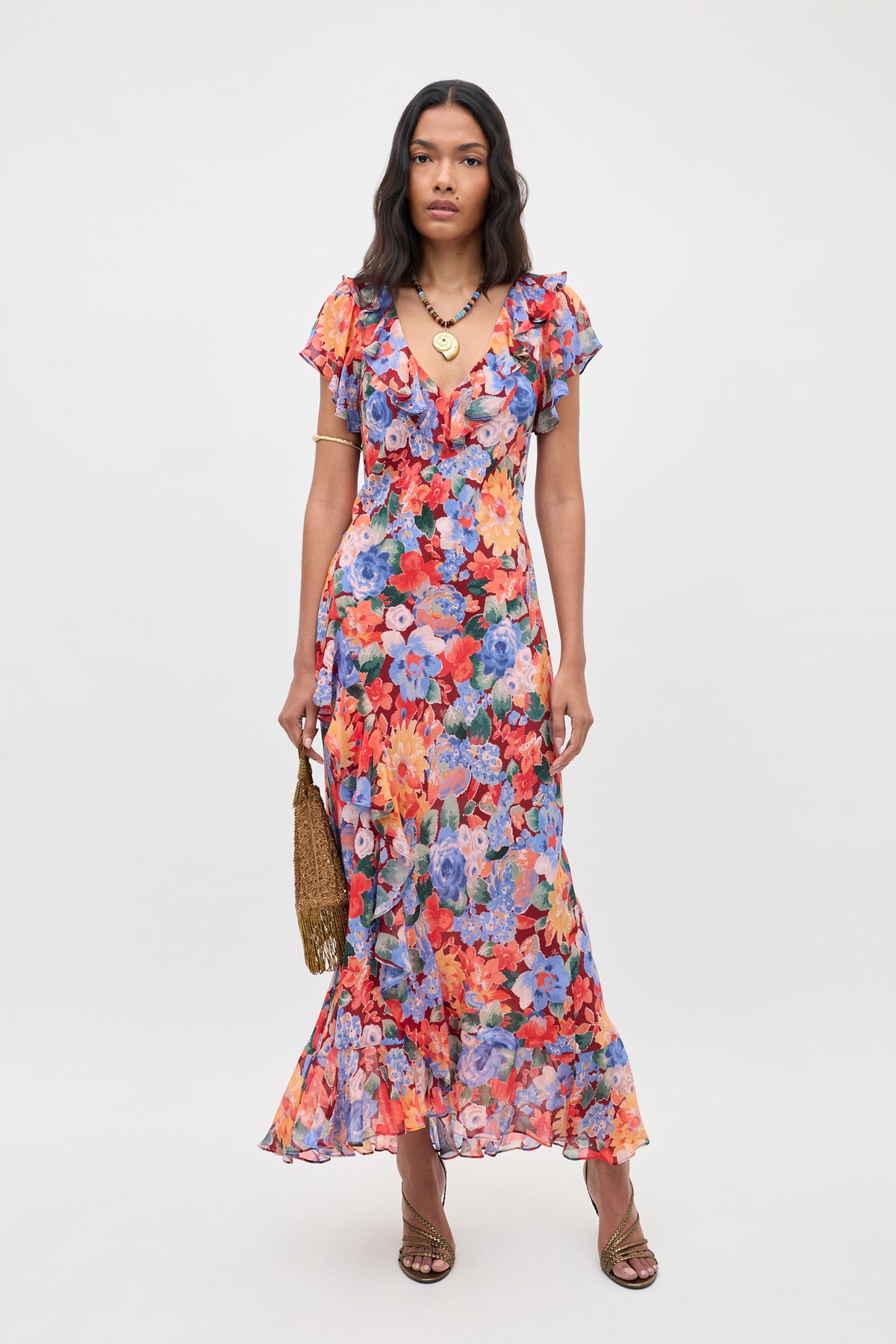 A woman wearing the Meadow - Chelsea Floral Red ankle-length dress, featuring a vibrant floral pattern, short sleeves, and ruffles, holds a small gold handbag and stands in strappy heels against a plain background.