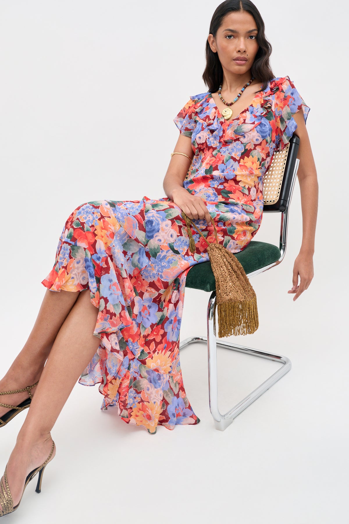 A woman in the Meadow - Chelsea Floral Red dress sits on a modern metal chair, holding a gold tasseled clutch. She wears gold heels and jewelry, with long dark hair against a plain white background.