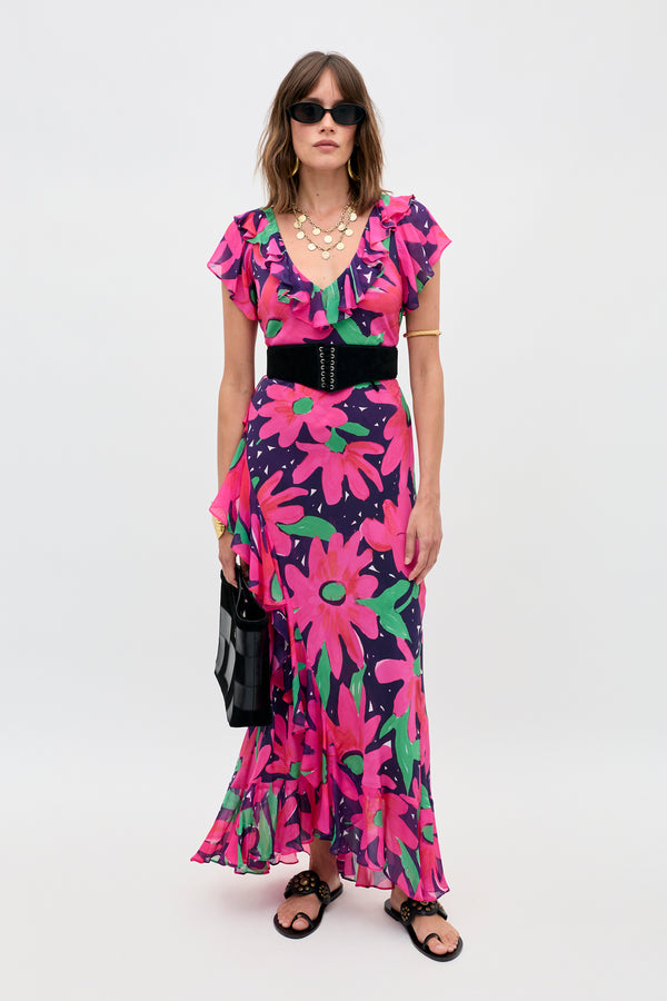 A woman wears the Meadow - Danish Floral Pink dress, a ruffled V-neck, floor-length style with a bold pink and green floral print. She pairs it with sunglasses, layered necklaces, a black belt, black sandals, and a black handbag against a plain background.
