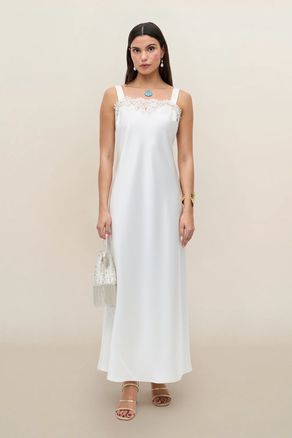 A woman with long dark hair wears the Maybelle - Ivory, a long white sleeveless bridal dress with lace details. She accessorizes with jewelry, sandals, and holds a beaded fringed handbag against a plain background.