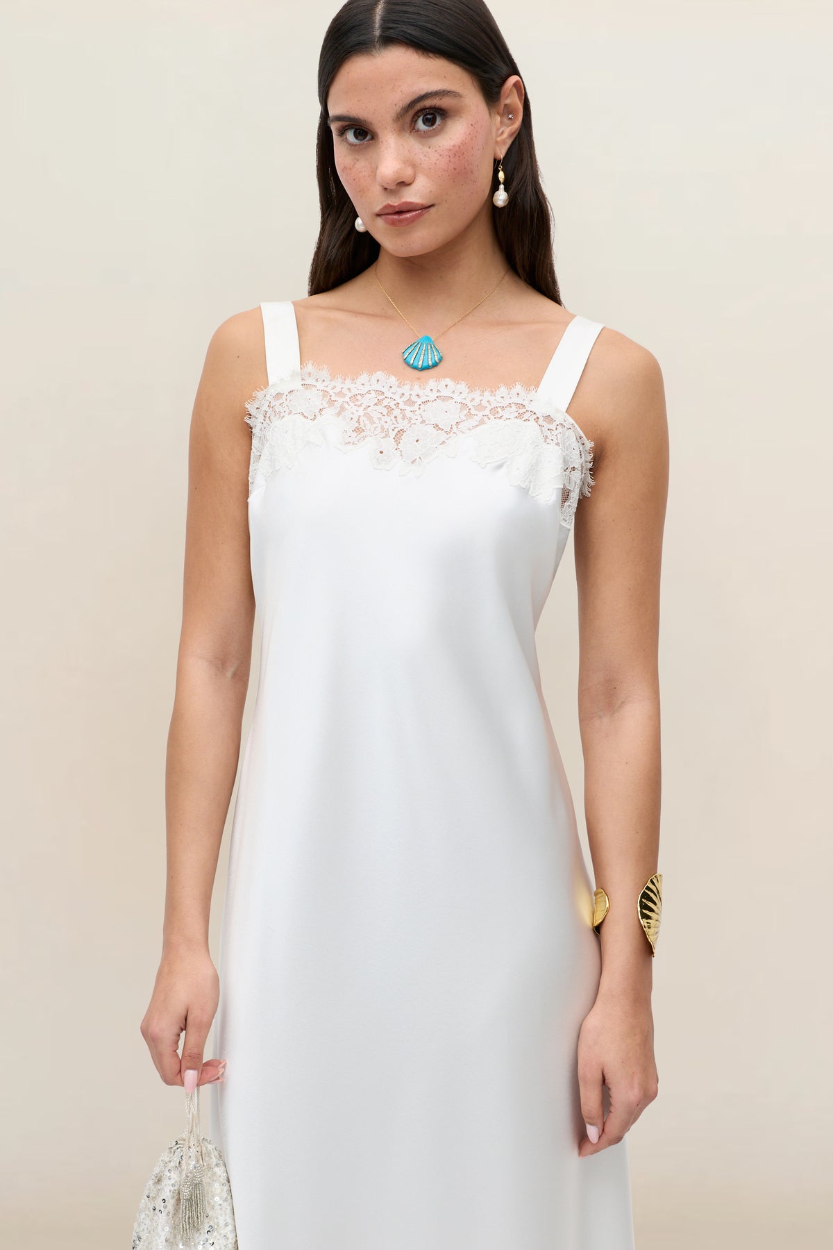 A woman wears the Maybelle - Ivory sleeveless bridal dress with lace details, holding a beaded bag. She has long brown hair, shell-shaped jewelry, and stands against a plain, light background.