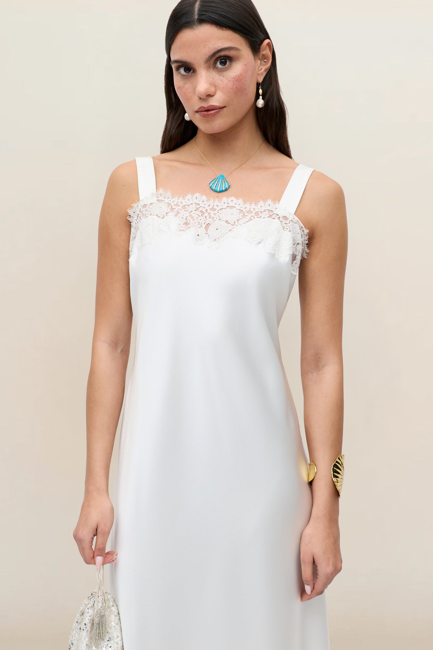 A woman wears the Maybelle - Ivory sleeveless bridal dress with lace details, holding a beaded bag. She has long brown hair, shell-shaped jewelry, and stands against a plain, light background.