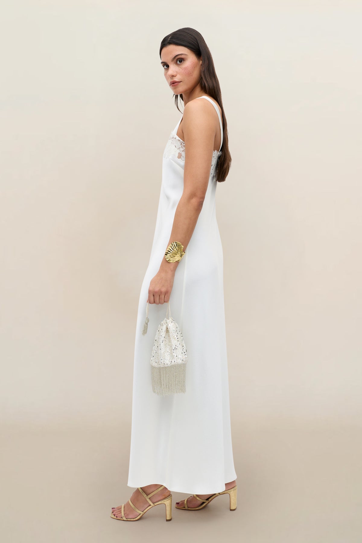 A woman with long dark hair stands sideways against a neutral background, wearing the Maybelle - Ivory bridal dress with lace detailing, gold strappy heels, a bold gold bracelet, and holding a beaded handbag.