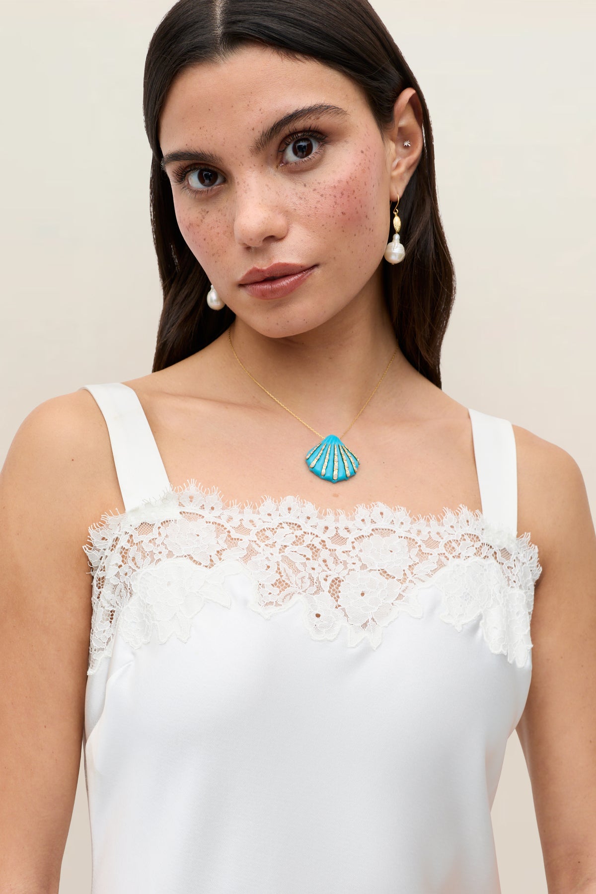A woman with long dark hair and freckles wears the Maybelle - Ivory satin bridal dress with lace details, pearl earrings, and a gold necklace with a blue shell pendant, standing against a plain light background.