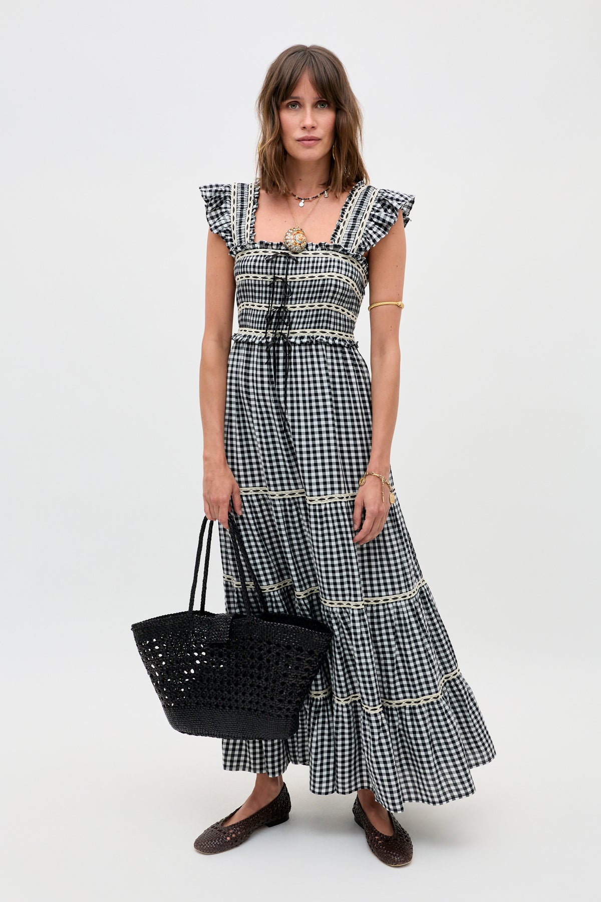 A woman stands against a plain background wearing the Chandra - Beth Check Black maxi dress with ruffled sleeves. She completes the look with a black woven tote, brown flats, layered necklaces, and an arm cuff.