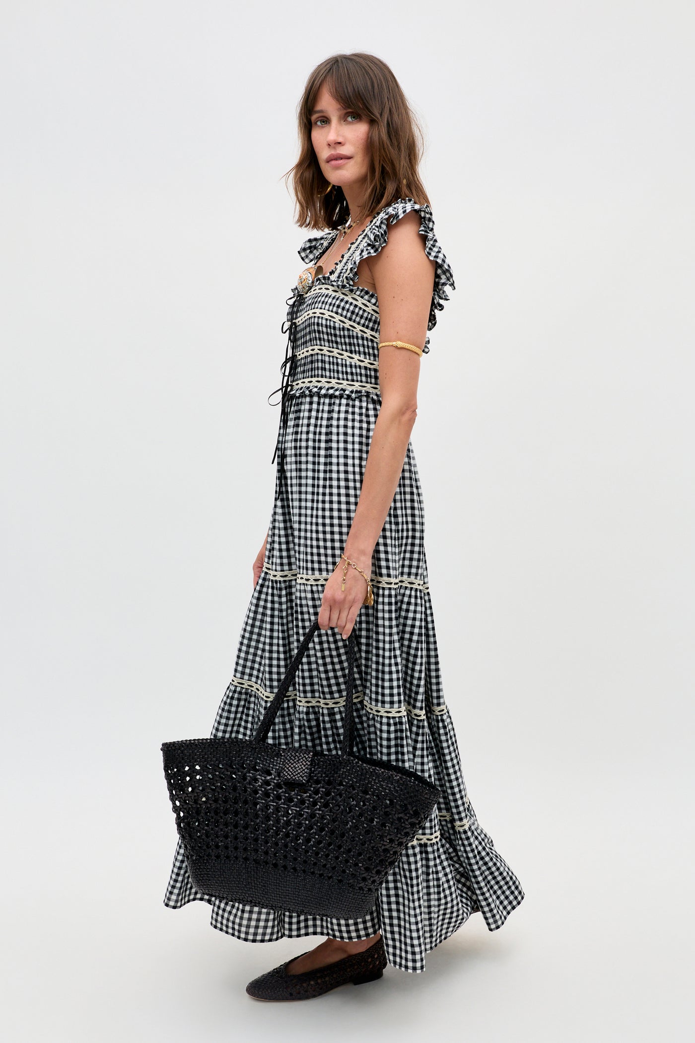A woman wears the Chandra - Beth Check Black maxi dress with ruffled sleeves, holding a black woven tote and wearing black flats, standing against a plain light background.