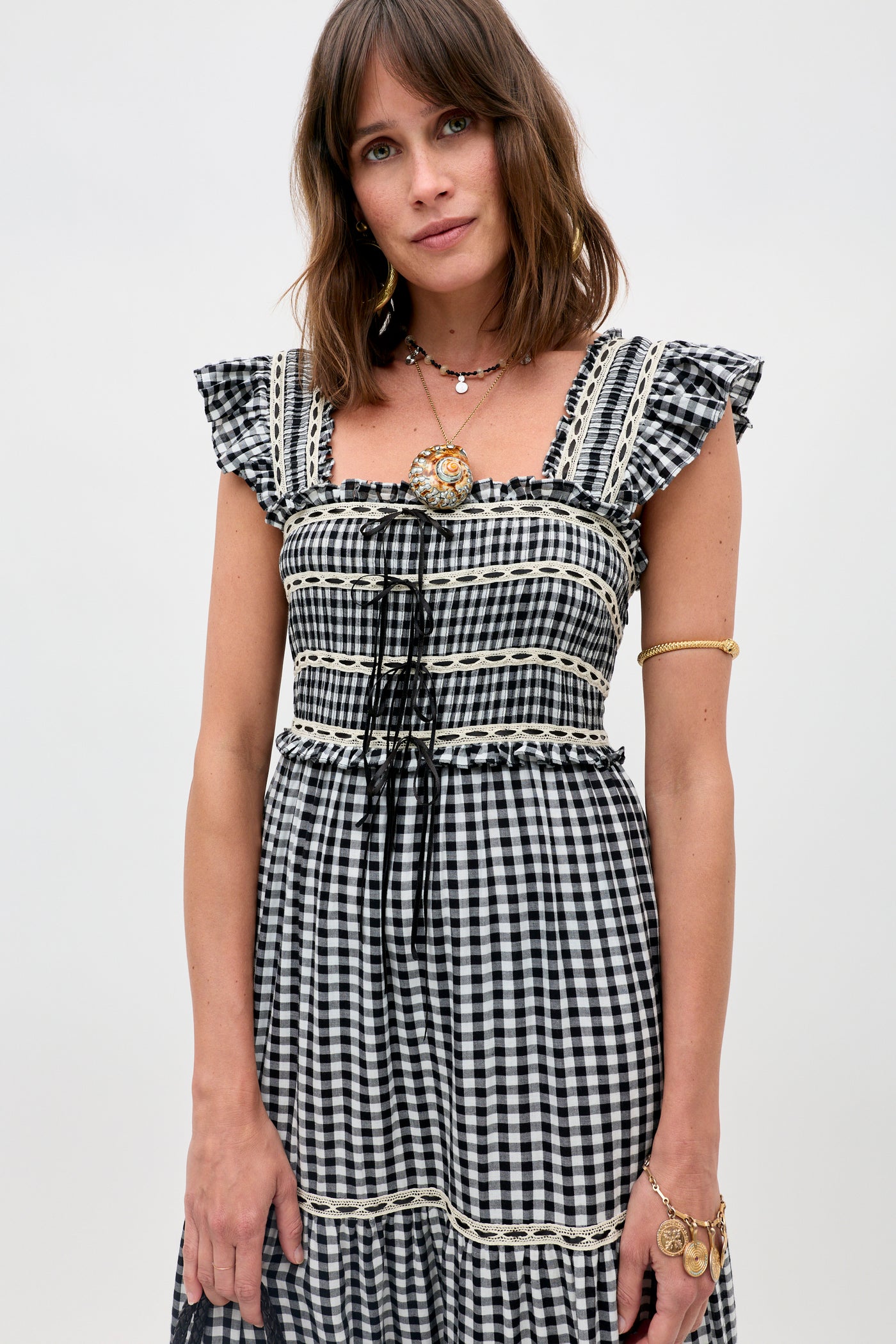 A woman with shoulder-length brown hair wears the Chandra - Beth Check Black dress, featuring ruffled sleeves and lace trim. She accessorizes with gold jewelry and a brooch, standing before a plain light background.