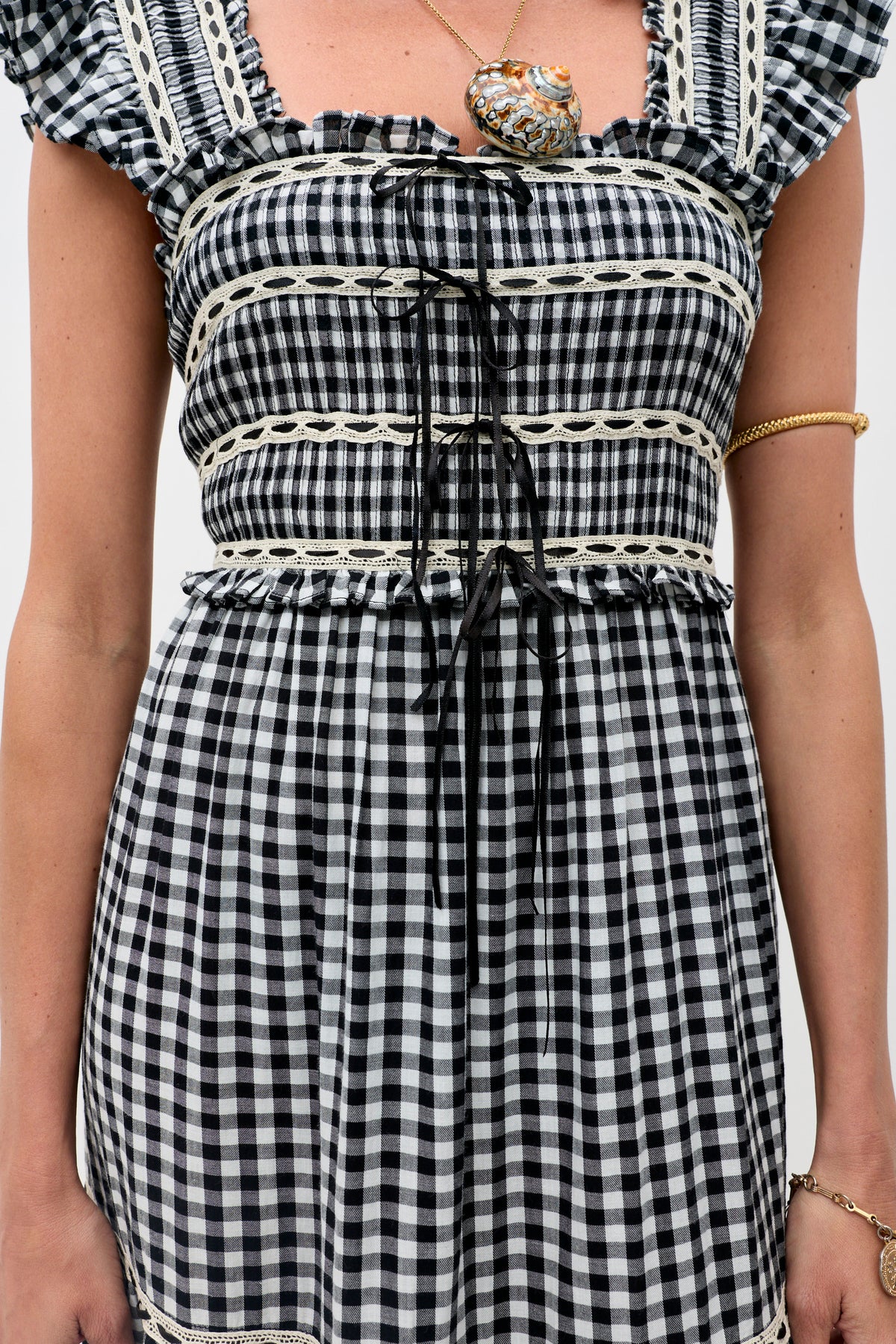A person wears the Chandra - Beth Check Black dress, featuring black and white gingham with lace trim and black ribbon ties. They accessorize with a gold bracelet and a large pendant necklace against a plain light background.