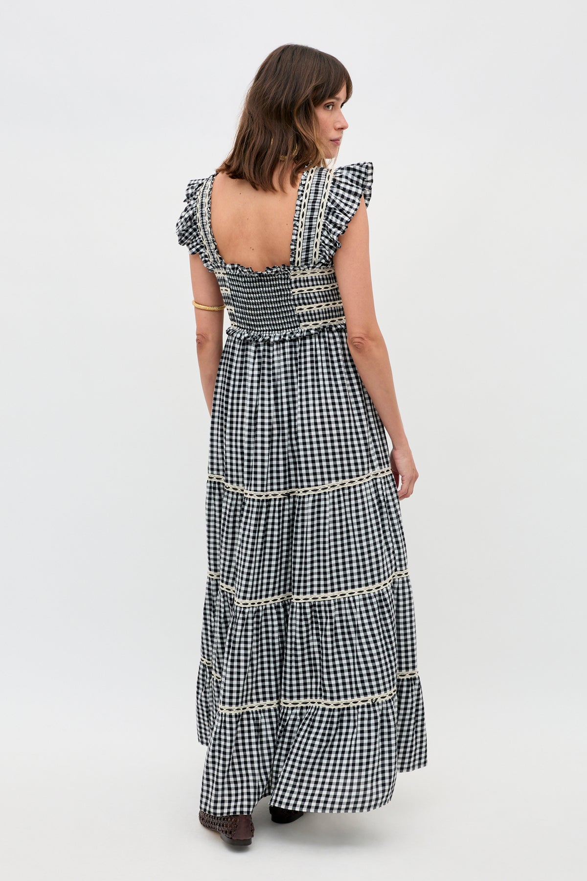 A woman with shoulder-length brown hair stands against a plain white background wearing the Chandra - Beth Check Black dress, featuring black-and-white gingham, ruffled sleeves, lace trim, a smocked bodice, and a tiered skirt.