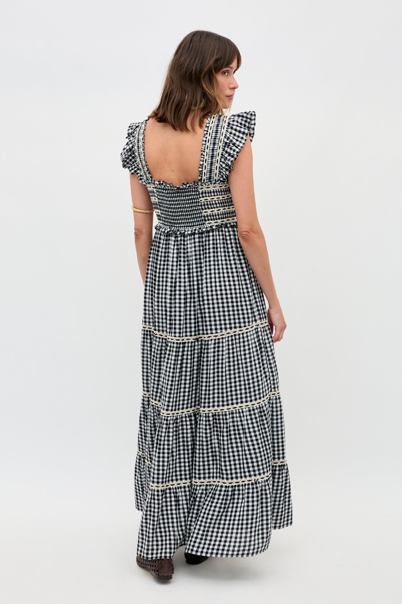 A woman with shoulder-length brown hair stands against a plain white background wearing the Chandra - Beth Check Black dress, featuring black-and-white gingham, ruffled sleeves, lace trim, a smocked bodice, and a tiered skirt.