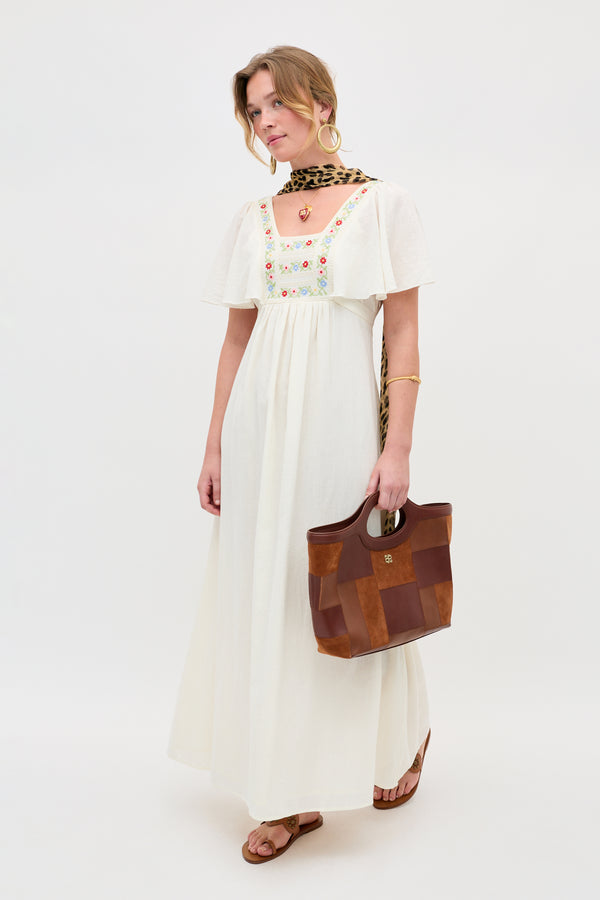 A woman is wearing the Jimena - Genoa Embroidery Cream long white dress with floral embroidery, paired with a leopard print scarf and hoop earrings. She holds a brown tote bag and wears sandals, standing against a plain white background.