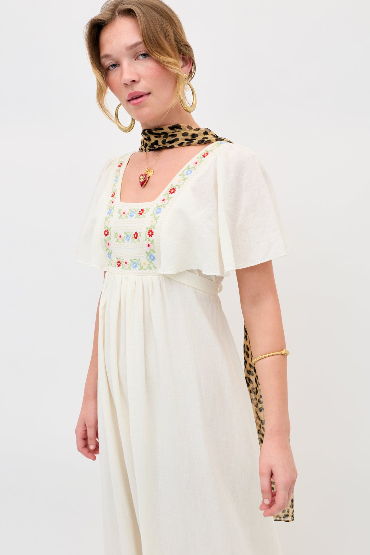 A woman wearing the Jimena - Genoa Embroidery Cream dress, paired with a leopard print scarf, gold hoop earrings, and a gold bracelet, standing against a plain light background.