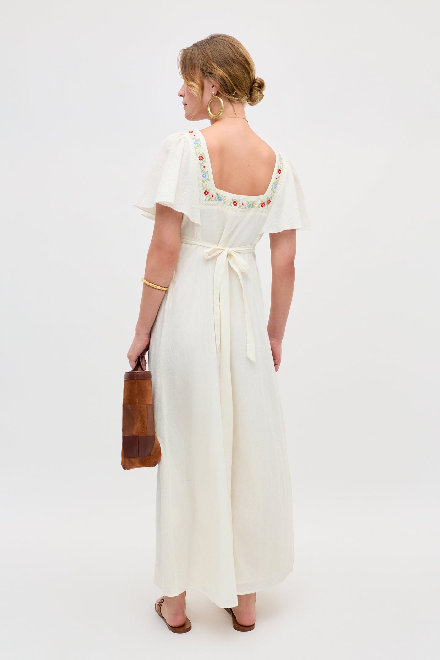 Woman in the Jimena - Genoa Embroidery Cream ankle-length dress with floral embroidery on the square neckline, tied at the waist, holding a brown bag, wearing large hoop earrings and sandals, standing against a plain white background.