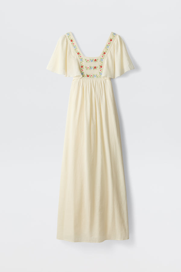 The Jimena - Genoa Embroidery Cream dress features a long, cream-colored design with flutter sleeves and vibrant floral embroidery accenting the square neckline and chest. Its flowing, relaxed silhouette and lightweight fabric create a summery, vintage-inspired look.