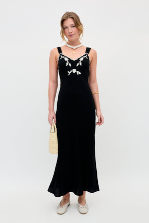 A woman poses before a plain backdrop in the Julietta - Tulipa Appliqué Black dress, featuring white floral embroidery at the neckline. She accessorizes with a woven bag, white shoes, gold jewelry, and her hair is in a loose updo.