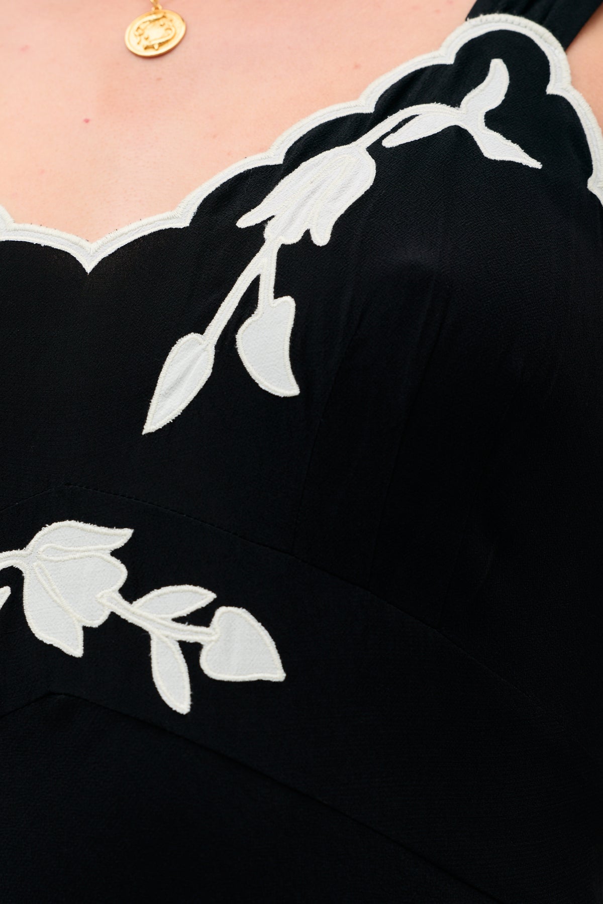 Close-up of a person wearing the Julietta - Tulipa Appliqué Black dress featuring white floral embroidery, scalloped edges, and a gold necklace with a round pendant.