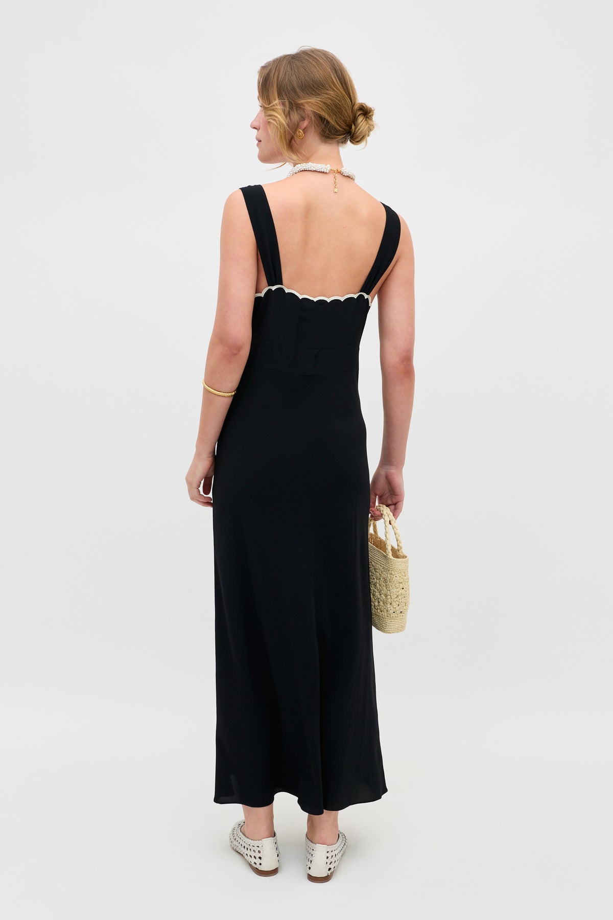 A woman faces away from the camera, standing against a plain backdrop, wearing the Julietta - Tulipa Appliqué Black sleeveless dress with scalloped edges, white slip-on shoes, a pearl necklace, and holding a woven handbag.