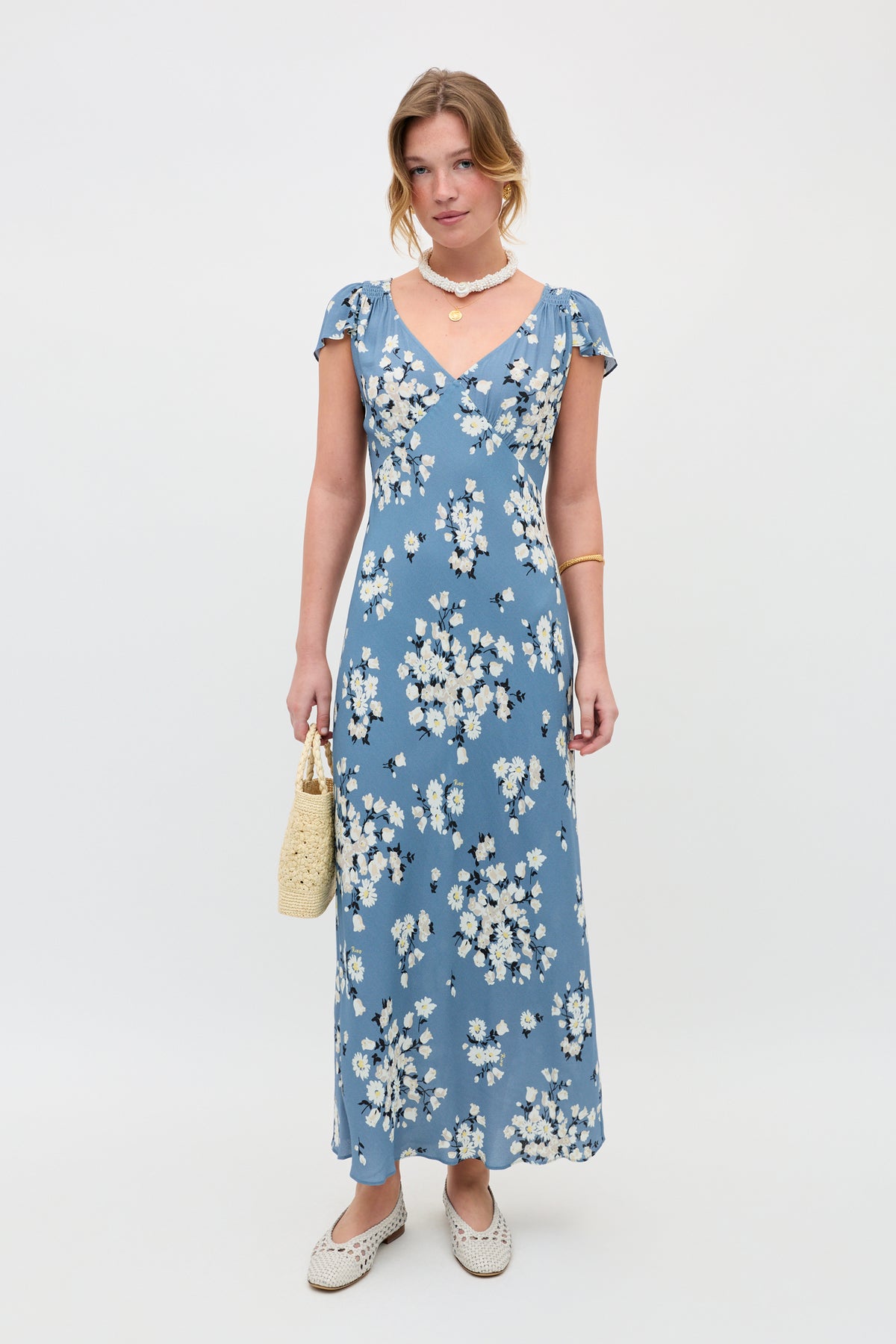 A woman stands against a plain background wearing the "Cathy - Amelia Bouquet Blue" dress, paired with white woven shoes, a beaded necklace, a gold bracelet, and holding a cream-colored woven handbag.