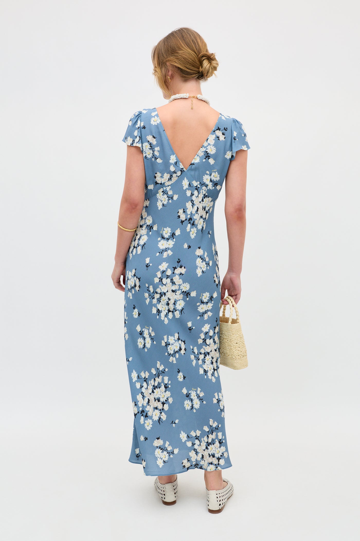 A woman, facing away, wears the Cathy - Amelia Bouquet Blue floral dress with a deep V-back, white shoes, and a woven handbag. Her hair is in a low bun and she accessorizes with a necklace and bracelet against a plain background.