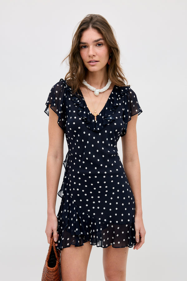 A woman wears the Tally - Layla Dot Navy dress, featuring white polka dots, ruffled sleeves, and a ruffled hem. She accessorizes with layered necklaces and a brown woven bag, standing against a plain light background.