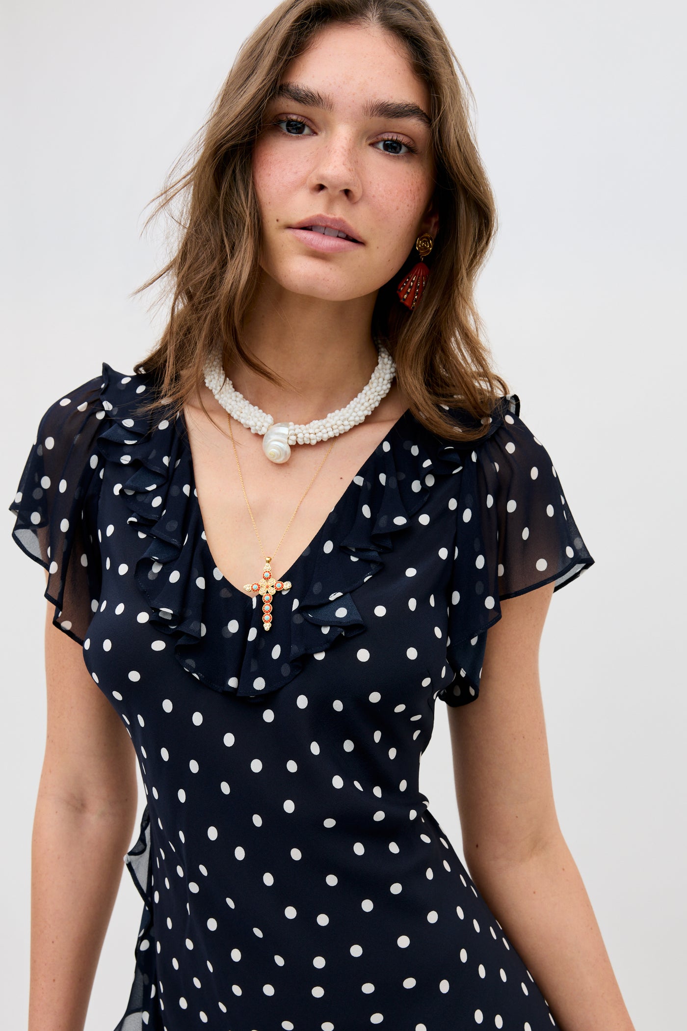 A woman with wavy brown hair wears the Tally - Layla Dot Navy dress, featuring white polka dots and ruffled sleeves. She pairs it with layered necklaces and red earrings, set against a plain light background.