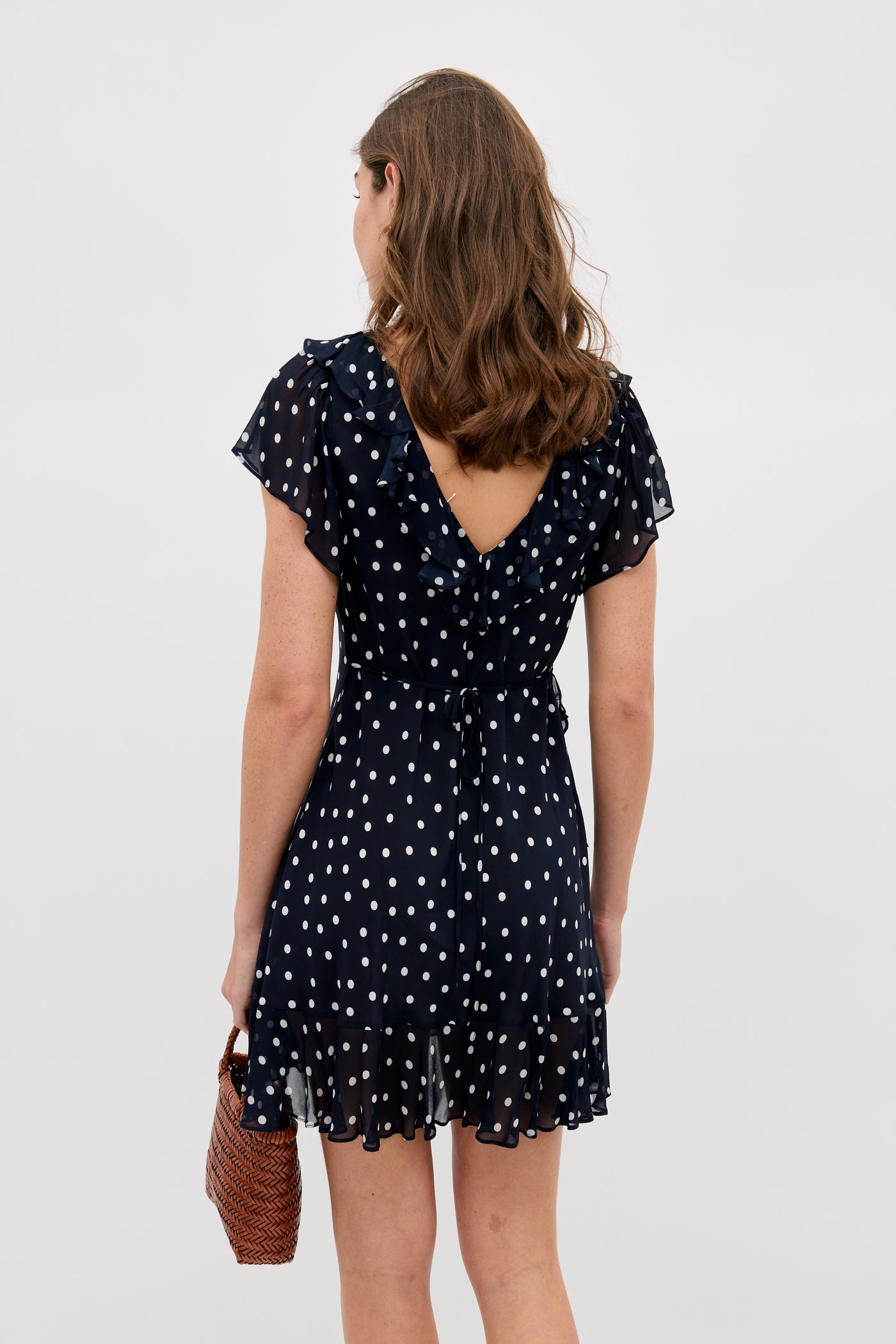 A woman with wavy brown hair, seen from behind, wears the Tally - Layla Dot Navy dress and holds a brown woven handbag against a plain light background.