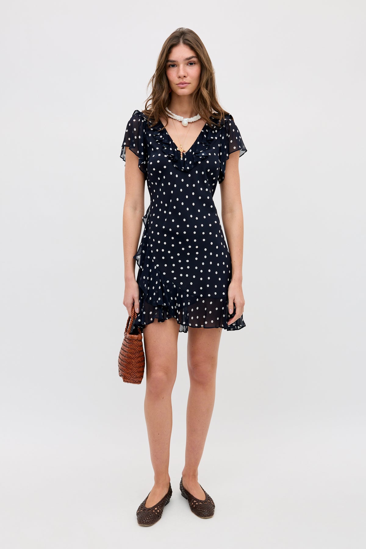 A woman stands against a plain background wearing the Tally - Layla Dot Navy dress, featuring white polka dots, ruffled sleeves and hem. She holds a brown woven handbag, wears dark flats, and accessorizes with a silver necklace.