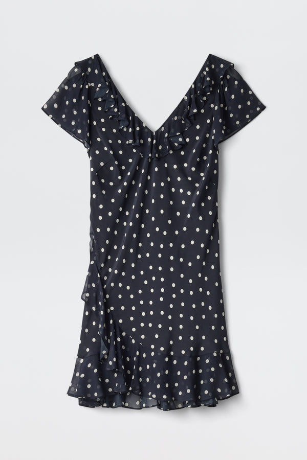 The Tally - Layla Dot Navy is a short-sleeved, navy blue dress with white polka dots, a V-neckline, ruffle details on the neckline and hem, and a slightly flared above-the-knee skirt.