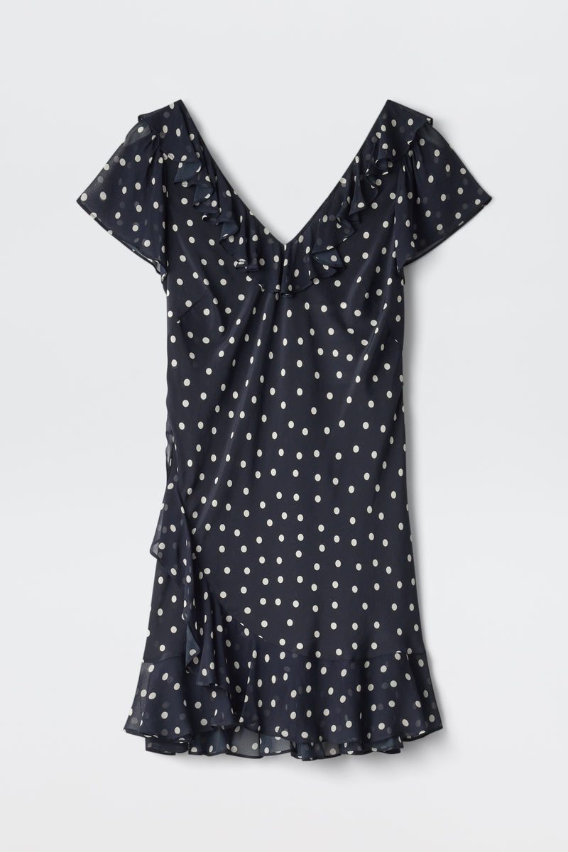 The Tally - Layla Dot Navy is a short-sleeved, navy blue dress with white polka dots, a V-neckline, ruffle details on the neckline and hem, and a slightly flared above-the-knee skirt.