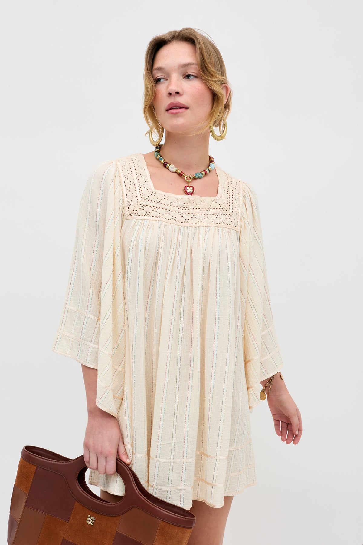 A woman with blonde hair wears the Estele - Cream dress, a loose-fitting cream-colored style with crochet detailing, paired with a colorful beaded necklace and a brown leather bag, standing against a plain light background.