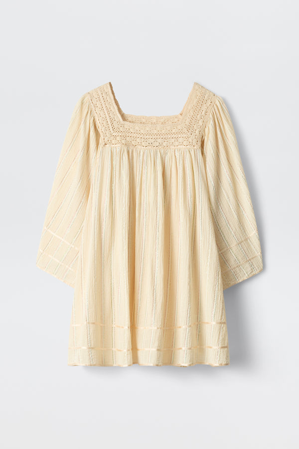 The Estele - Cream is a cream-colored, long-sleeve toddler top featuring a textured, square neckline and subtle vertical stripes. It offers a loose fit with decorative stitching on the chest, sleeves, and hem.
