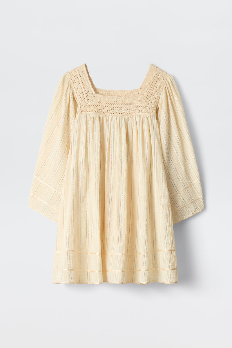 The Estele - Cream is a cream-colored, long-sleeve toddler top featuring a textured, square neckline and subtle vertical stripes. It offers a loose fit with decorative stitching on the chest, sleeves, and hem.