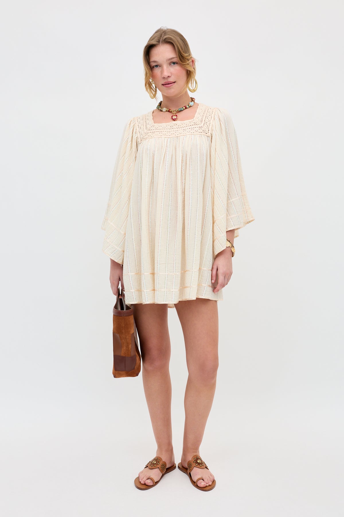 A woman stands against a white background wearing the Estele - Cream, a loose, long-sleeve dress, paired with brown sandals, a choker necklace, bracelets, and holding a brown handbag.